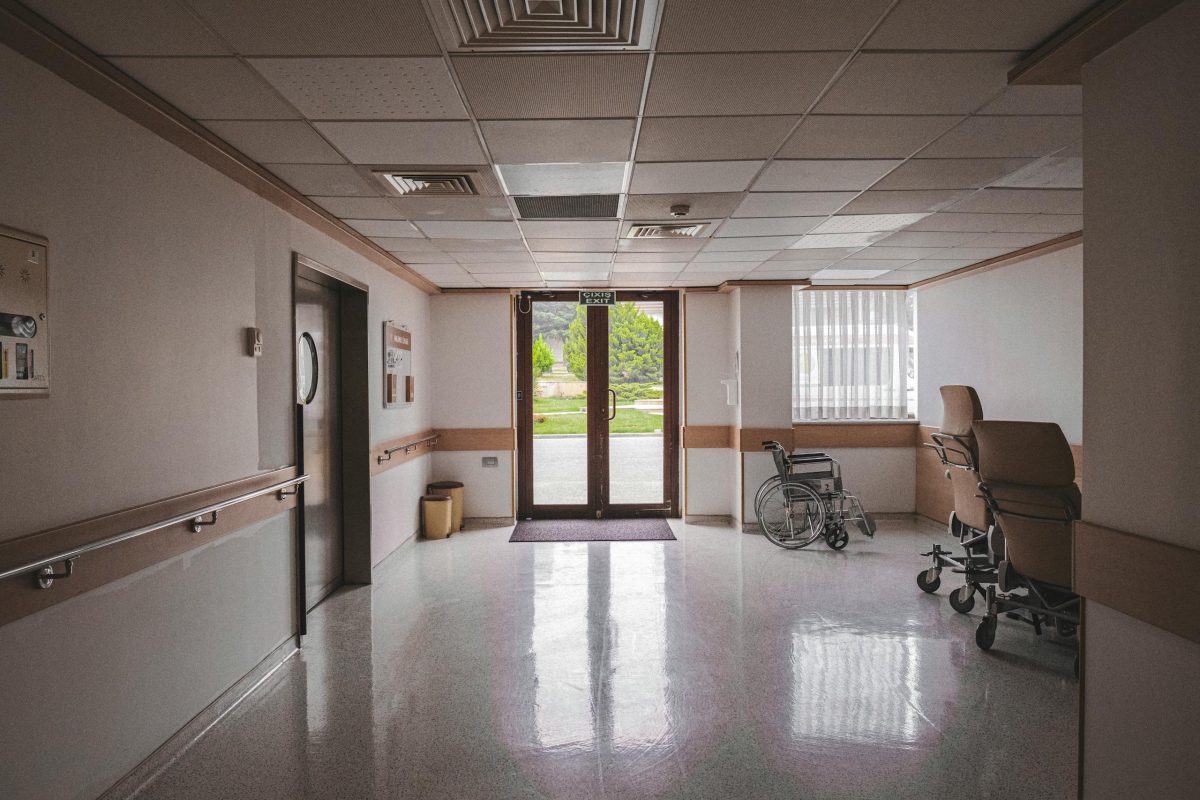 Massage en fin de vie imminente 7 Hospital corridor with wheelchair, reflecting glass doors, and outdoor view. Ideal for healthcare themes.